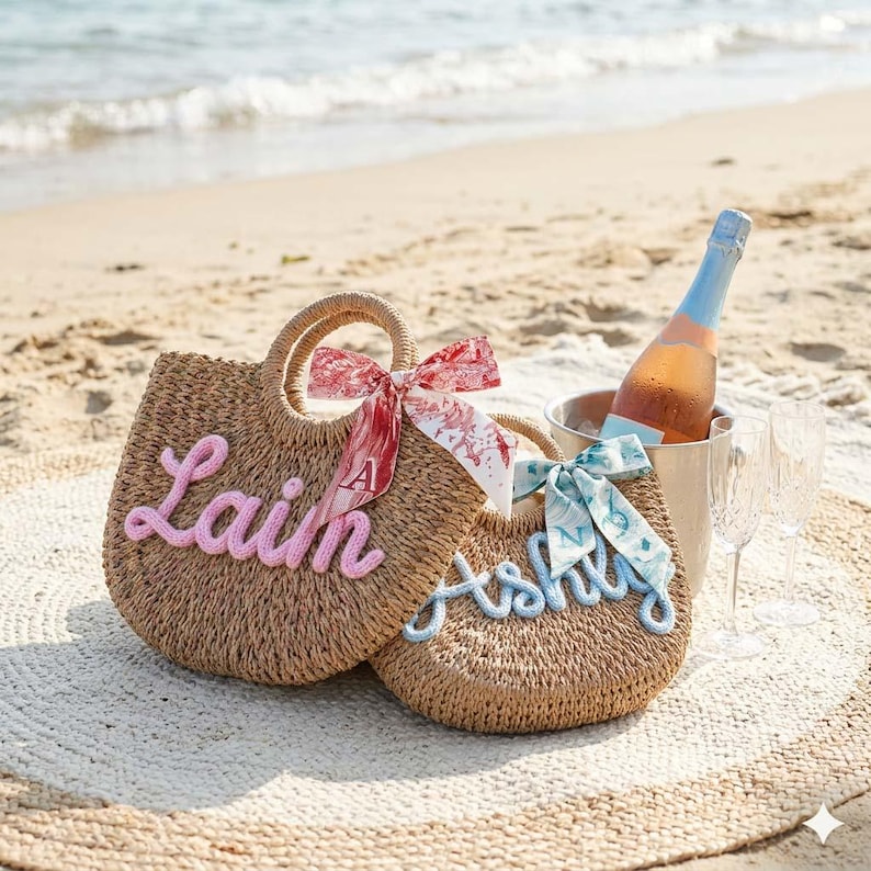 May include: Two woven straw tote bags with handles, personalized with the names "Lain" in pink and "Ashly" in blue. Each bag has a decorative ribbon. A bottle of ros&eacute; in an ice bucket and two champagne flutes sit on a beach blanket.
