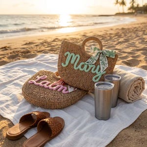 May include: Two woven straw tote bags with the names "Mary" and "Lain" embroidered in green and pink, respectively. The bags are on a white blanket with two stainless steel tumblers, a rolled-up beige towel, and brown leather sandals. The scene is set on a sandy beach.