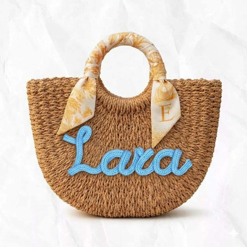 May include: A woven straw tote bag with a rounded shape. The bag features a light blue knitted name "Lara" on the front. The handles are wrapped with a cream-colored scarf with a gold pattern. The bag is designed for carrying items.