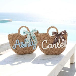 May include: Two woven straw tote bags with rounded handles. One bag has the name "Ashly" in light blue script, and the other has "Carle" in white script. Both bags have decorative bows. The background shows a pool and the ocean.