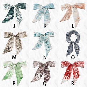 May include: Collection of silk scarves with various designs and initial letters. The scarves feature floral, map, and abstract patterns in colors like green, blue, brown, and red. Each scarf has a bow shape with long, flowing ends.