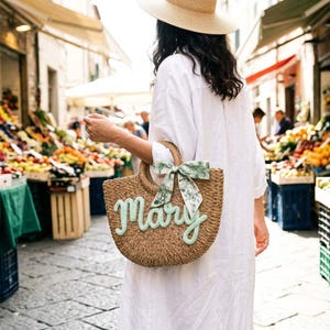 May include: A woven straw tote bag with the name "Mary" in light green script. The bag has a rounded shape with a handle and a decorative green and white patterned bow. The background shows a market scene.