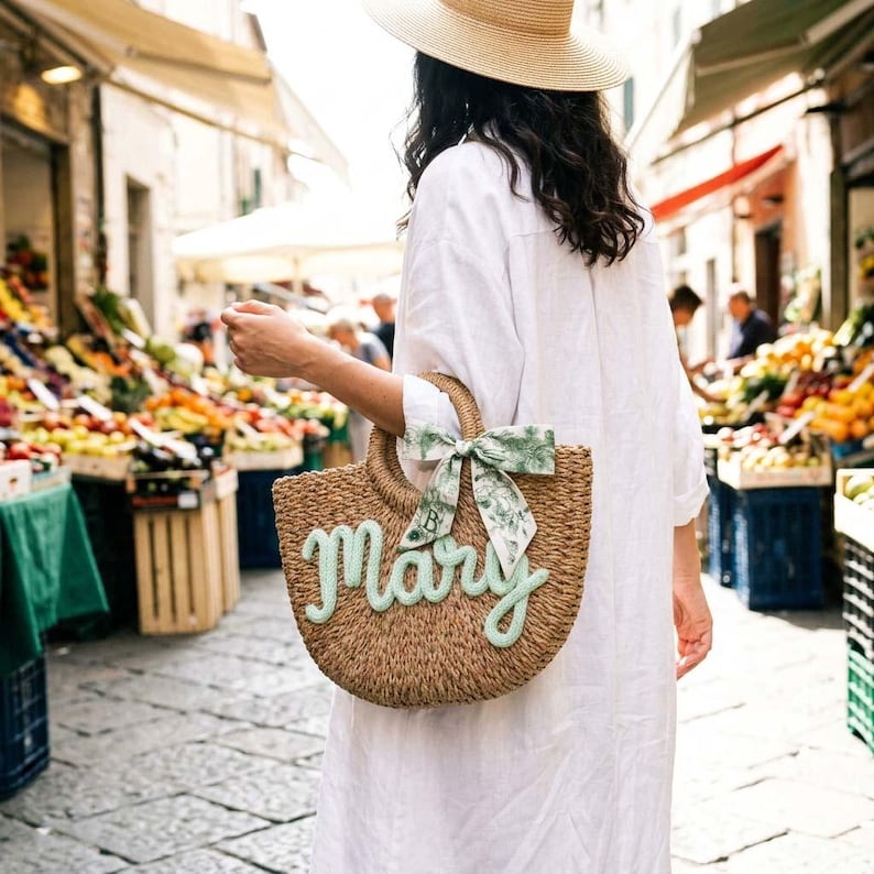 May include: A woven straw tote bag with the name "Mary" in light green script. The bag has a rounded shape with a handle and a decorative green and white patterned bow. The background shows a market scene.