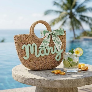 May include: A woven straw tote bag with the name "Mary" in light green script. The bag has a round handle and a green and white patterned bow. A cocktail with lime and orange slices sits nearby on a stone table.