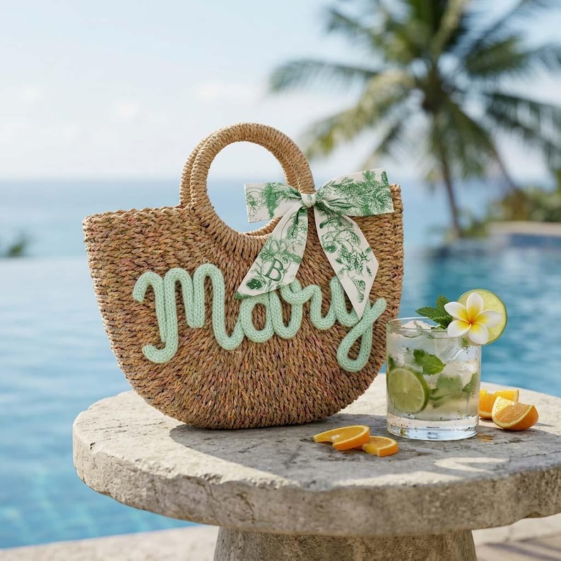 May include: A woven straw tote bag with the name "Mary" in light green script. The bag has a round handle and a green and white patterned bow. A cocktail with lime and orange slices sits nearby on a stone table.
