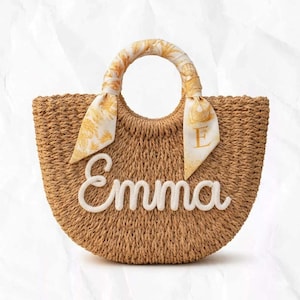 May include: A woven straw tote bag with a rounded shape. The bag features a handle wrapped with a patterned scarf in shades of yellow and white. The name "Emma" is spelled out in white cursive letters on the front.