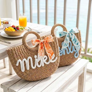 May include: Two woven straw tote bags with round handles. One bag has the name "Mike" in white script, and an orange and white patterned bow. The other bag has the name "Emily" in blue script, and a blue and white patterned bow.