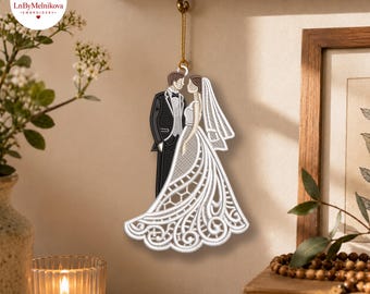 May include: A hanging embroidered ornament depicting a couple in wedding attire. The figure on the left wears a black suit with a white shirt and bow tie. The figure on the right wears a white lace-patterned gown and a matching veil.