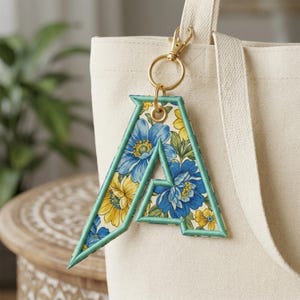 May include: A letter A keychain featuring a floral pattern with blue and yellow flowers on a cream background, bordered by teal stitching. The letter is attached to a gold-toned metal lobster clasp and ring, hanging from a beige canvas tote bag.