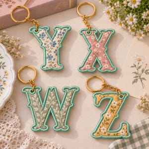Könnte beinhalten: Vier bestickte Buchstaben-Schlüsselanhänger mit den Buchstaben Y, X, W und Z. Jeder Buchstabe hat ein florales Muster in Blau, Rosa, Grün oder Gelb, einen mintgrünen gestickten Rand und einen goldfarbenen Metallring an einer Kette.