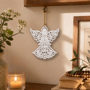 May include: A white, intricately designed angel ornament with detailed cut-out patterns. The angel is suspended by a gold string, showcasing its delicate wings and flowing gown. The ornament is hanging from a wooden shelf, adding a touch of elegance.