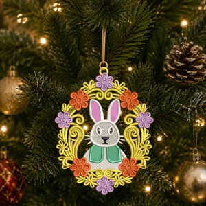 May include: Embroidered Easter bunny ornament with a yellow floral frame, orange and purple flowers, and a gold hanging string. The bunny has pink ears, a gray face, and a green vest. The ornament is hanging on a Christmas tree.