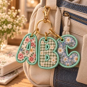 Puede incluir: Un llavero de metal dorado con tres letras de tela acolchada, A, B y C, unidas a una anilla central. La letra A tiene un estampado floral rosa, la B un patrón de cuadros vichy verdes con margaritas y la C un estampado floral azul, con bordes cosidos en verde menta.