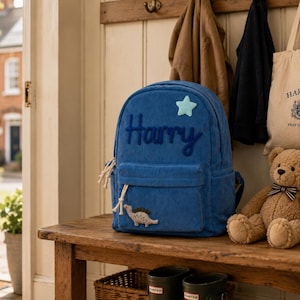 May include: A blue corduroy backpack featuring the name Harry embroidered in dark blue thread, a light blue star patch, and a small dinosaur patch on the front pocket. The backpack has a zipper closure with white cord pulls and side pockets.