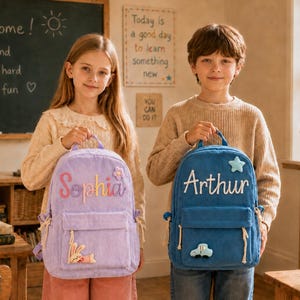 Puede incluir: Una mochila de pana color lavanda con el nombre Sophia bordado en rosa y parches de flor y conejo, y una mochila de pana azul con el nombre Arthur bordado en blanco y parches de estrella y coche, ambas con bolsillos frontales.