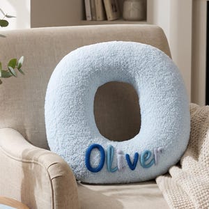 May include: A light blue, textured, circular-shaped cushion featuring the name Oliver embroidered in a mix of dark blue, medium blue, and light grey fabric letters across the bottom front section of the plush, soft-touch material.
