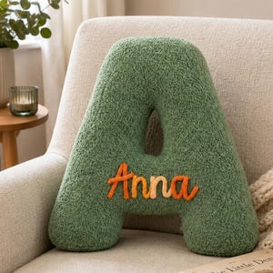 May include: A sage green letter A-shaped throw cushion made of textured sherpa fabric. The front of the cushion features the name Anna embroidered in a gradient of orange and yellow thread across the centre of the letter shape.