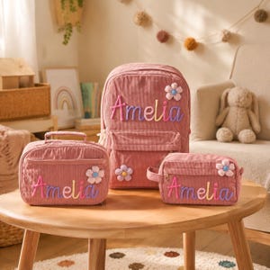 Peut inclure: Un sac à dos, un sac à déjeuner rectangulaire et une trousse en velours côtelé rose poudré, chacun portant le prénom Amelia brodé en lettres colorées et une petite fleur en tissu. L'ensemble est en velours côtelé texturé avec fermetures éclair.