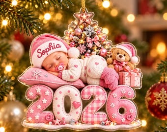 Peut inclure: Une décoration de Noël en forme de sapin représentant un bébé endormi portant un bonnet rose avec le prénom Sophia, un ours en peluche et un cadeau. La base affiche l'année 2026 en grands chiffres roses à motifs. Inclut une étoile au sommet.