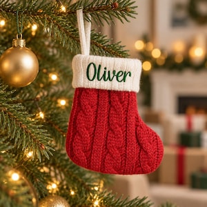 May include: A small red cable-knit Christmas stocking featuring a white ribbed cuff with the name Oliver embroidered in green script. The stocking includes a white hanging loop and is displayed hanging from a green pine tree branch.