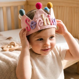 May include: A light pink fabric crown featuring the name Sophia embroidered in multi-colored yarn letters. The crown is decorated with small, round, colorful pom-poms attached to each of the five pointed peaks along the top edge of the band.