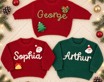 May include: Three long-sleeve knit sweaters in red and green colors. The top red sweater features the name George in gold script with a gift and tree patch. The bottom red sweater says Sophia with a Santa and bell patch. The green sweater says Arthur.