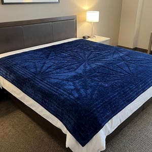 May include: A deep navy blue velvet blanket featuring a geometric starburst pattern with stitched lines. The blanket is draped over a bed with a white sheet and a dark brown headboard, creating a textured surface with a soft, plush appearance.