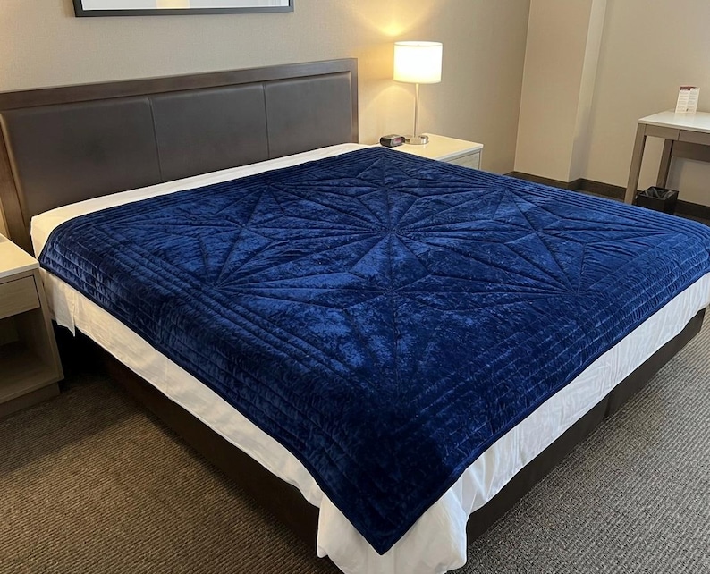 May include: A deep navy blue velvet blanket featuring a geometric starburst pattern with stitched lines. The blanket is draped over a bed with a white sheet and a dark brown headboard, creating a textured surface with a soft, plush appearance.