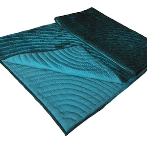 May include: A rectangular teal velvet quilt featuring a stitched pattern of concentric semicircles. The quilt is folded over to reveal a lighter teal underside with matching stitching. The fabric has a soft, reflective sheen throughout.
