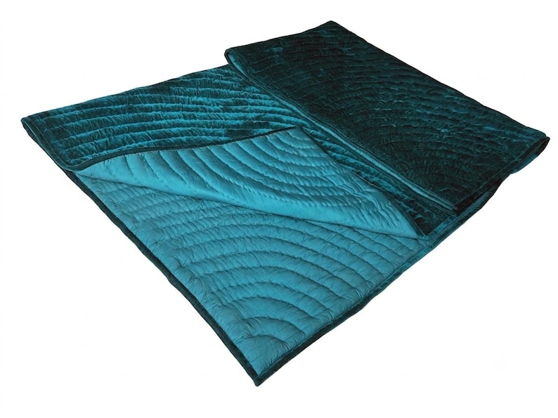 May include: A rectangular teal velvet quilt featuring a stitched pattern of concentric semicircles. The quilt is folded over to reveal a lighter teal underside with matching stitching. The fabric has a soft, reflective sheen throughout.