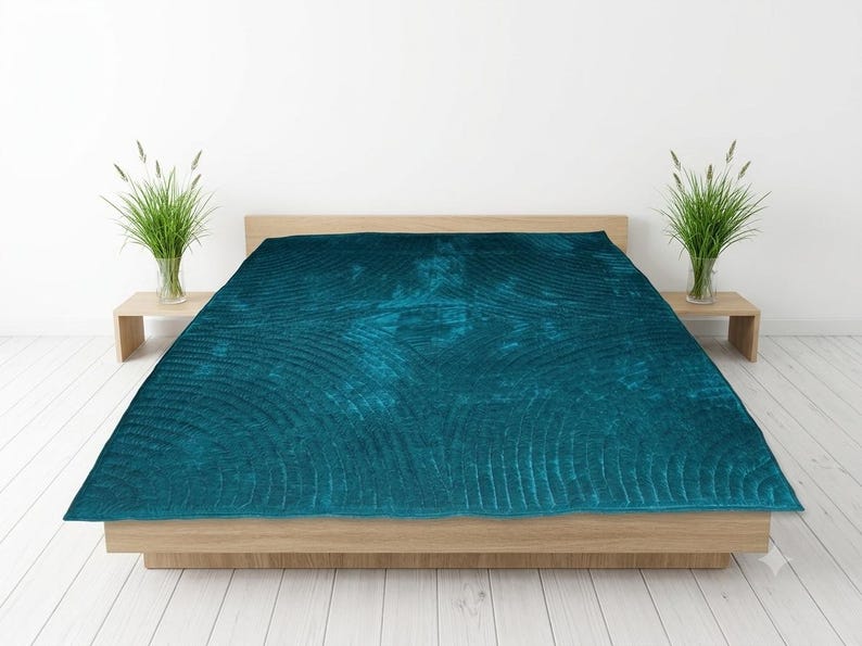 May include: A deep teal velvet quilt featuring a textured, stitched pattern of concentric semicircles. The quilt is draped over a low-profile wooden bed frame, creating a rich, saturated focal point with its dark blue-green color and soft sheen.