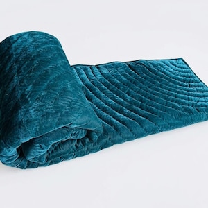 May include: A deep teal velvet quilt featuring a soft, plush texture and a series of parallel, curved quilted lines that create a subtle wave pattern across the fabric. The quilt is partially rolled up, showcasing its thick, padded construction.