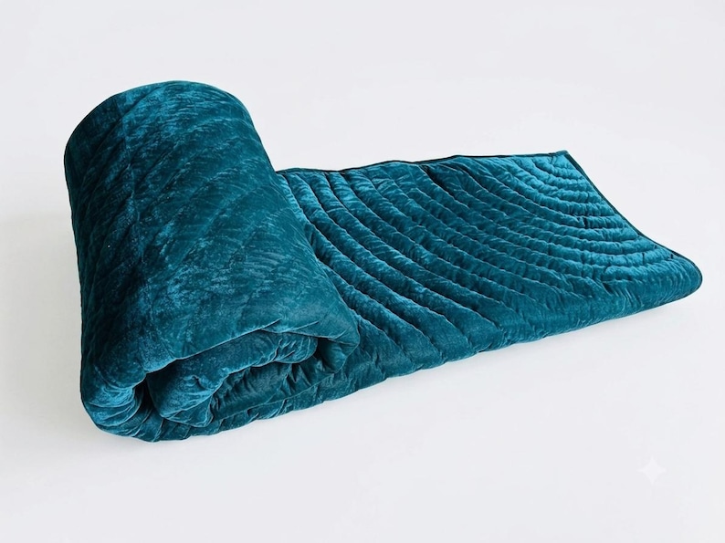 May include: A deep teal velvet quilt featuring a soft, plush texture and a series of parallel, curved quilted lines that create a subtle wave pattern across the fabric. The quilt is partially rolled up, showcasing its thick, padded construction.