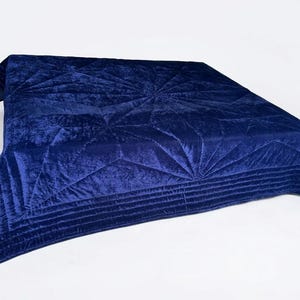 May include: A deep navy blue velvet quilt featuring a geometric starburst pattern created by intricate stitching. The fabric has a soft, reflective sheen, and the edges are finished with a wide border containing multiple parallel horizontal lines.