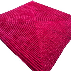 Puede incluir: Una manta fucsia vibrante con un dise&ntilde;o acolchado. La manta presenta una serie de l&iacute;neas paralelas que crean una superficie texturizada. Los bordes est&aacute;n acabados con un patr&oacute;n contrastante, agregando inter&eacute;s visual. La manta es cuadrada.