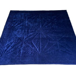 May include: A deep navy blue velvet quilt featuring a geometric star pattern created by intricate stitching. The square textile has a soft, reflective surface with a wide border composed of multiple parallel quilted lines framing the central design.