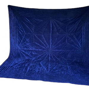May include: A deep royal blue velvet quilt featuring a geometric starburst pattern created by intricate stitching. The rectangular textile has a smooth, lustrous surface with multiple parallel stitched lines forming a border around the central design.