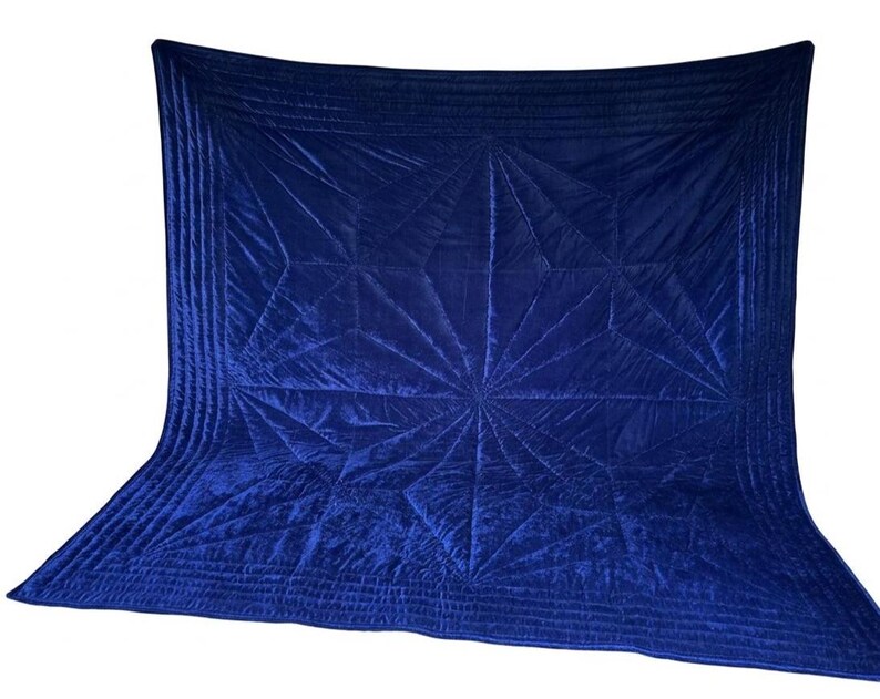 May include: A deep royal blue velvet quilt featuring a geometric starburst pattern created by intricate stitching. The rectangular textile has a smooth, lustrous surface with multiple parallel stitched lines forming a border around the central design.