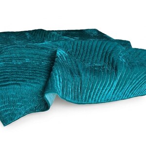 May include: A teal velvet quilt featuring a series of horizontal channel stitching across the entire surface. The fabric has a soft, reflective sheen and is folded over itself, displaying a consistent deep blue-green color throughout.