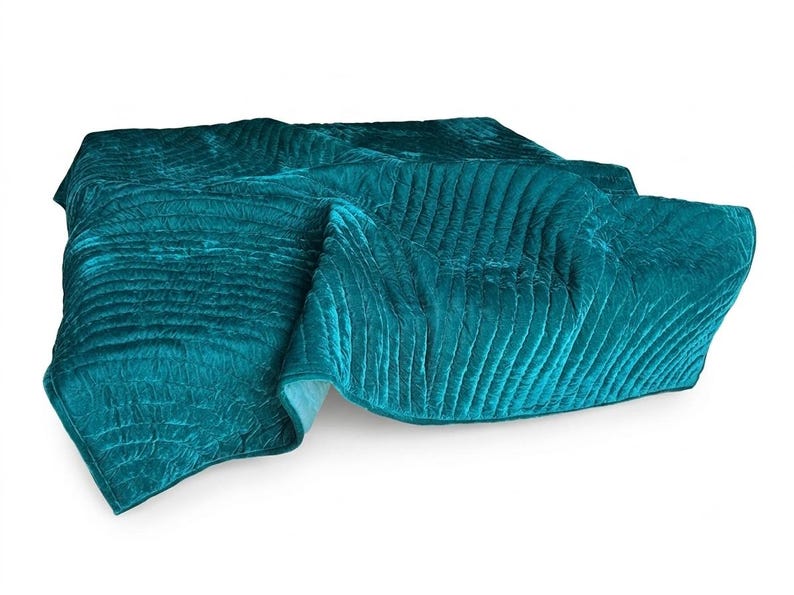 May include: A teal velvet quilt featuring a series of horizontal channel stitching across the entire surface. The fabric has a soft, reflective sheen and is folded over itself, displaying a consistent deep blue-green color throughout.
