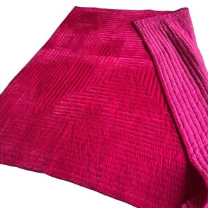 Puede incluir: Una vibrante manta acolchada en tonos fucsia y magenta. La manta presenta un patr&oacute;n geom&eacute;trico en el cuerpo principal y un borde de color liso contrastante. La tela tiene una textura suave y aterciopelada, lo que sugiere comodidad y calidez.