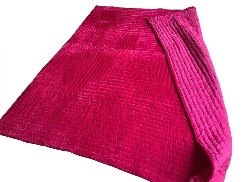 Puede incluir: Una vibrante manta acolchada en tonos fucsia y magenta. La manta presenta un patr&oacute;n geom&eacute;trico en el cuerpo principal y un borde de color liso contrastante. La tela tiene una textura suave y aterciopelada, lo que sugiere comodidad y calidez.