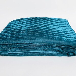 May include: A folded teal velvet quilt featuring a series of diagonal quilted channels across the surface. The fabric has a soft, reflective sheen characteristic of velvet, with deep blue-green tones and visible stitching along the quilted lines.