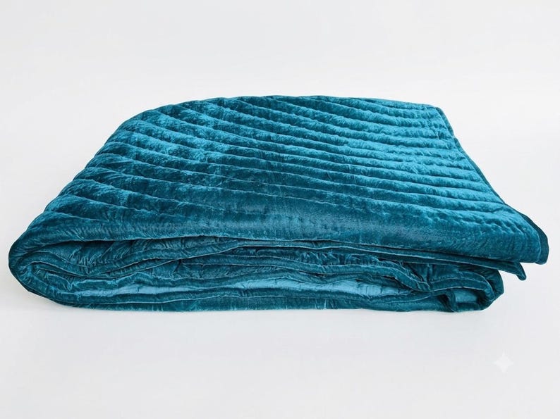 May include: A folded teal velvet quilt featuring a series of diagonal quilted channels across the surface. The fabric has a soft, reflective sheen characteristic of velvet, with deep blue-green tones and visible stitching along the quilted lines.