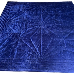 May include: A square, deep blue velvet quilt featuring a central geometric star pattern created by intricate stitching. The border consists of multiple parallel quilted lines that frame the central design, providing a textured appearance.