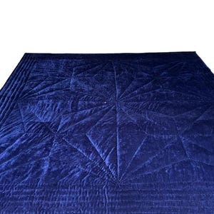 May include: A deep navy blue velvet quilt featuring a prominent geometric star pattern created by intricate stitching. The fabric has a soft, reflective sheen, and the border is finished with multiple rows of parallel straight-line quilting.