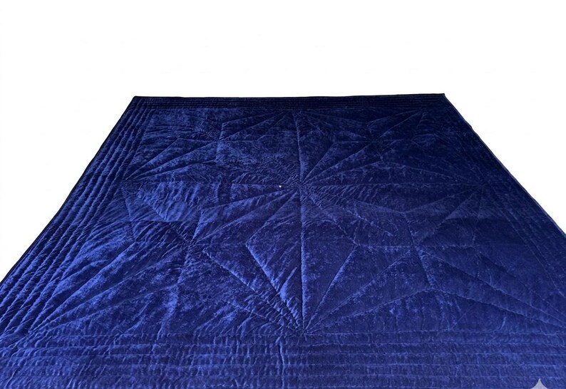 May include: A deep navy blue velvet quilt featuring a prominent geometric star pattern created by intricate stitching. The fabric has a soft, reflective sheen, and the border is finished with multiple rows of parallel straight-line quilting.