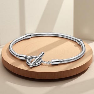 May include: A silver snake chain bracelet featuring a toggle clasp closure. The bracelet has a circular shape with two fixed stations along the chain and a central heart-shaped ring with a bar that slides through to secure the jewellery piece.