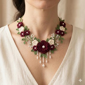 May include: A handcrafted floral necklace featuring burgundy and cream-coloured crochet flowers with green leaves and pearl accents. The necklace has a delicate, feminine design, with dangling pearl strands.
