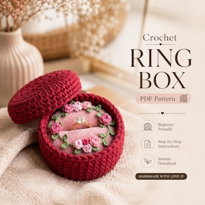 May include: A deep red crocheted ring box with a matching lid. Inside, a pink velvet lining and crocheted flowers surround a gold ring with a diamond. The image includes text that reads "Crochet Ring Box PDF Pattern".